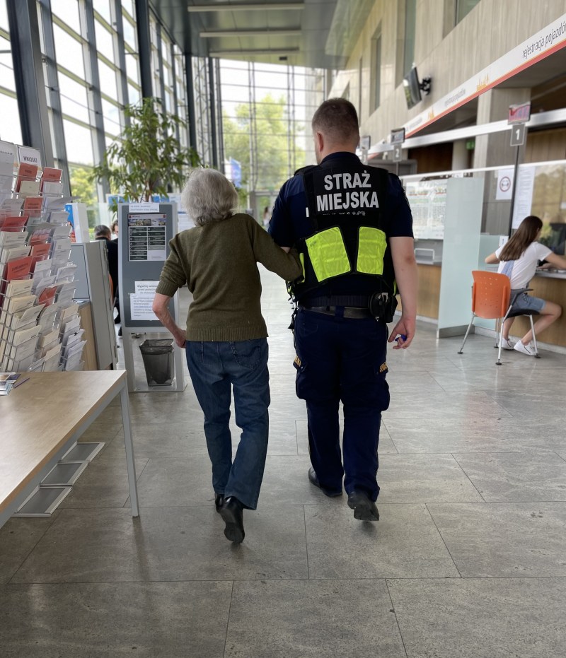 Strażnik miejski prowadzący pod ramię starszą kobietę na korytarzu urzędu dzielnicy w Wilanowie.