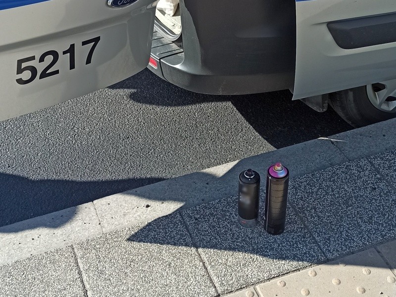 Dwie puszki z farbą w aerozolu stoją na chodniku obok radiowozu Straży Miejskiej.