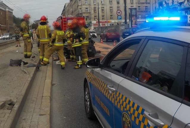 Radiowóz straży miejskiej zabezpiecza miejsce wypadku samochodowego. Na pierwszym planie radiowóz, dalej strażacy przy zniszczonym pojeździe.