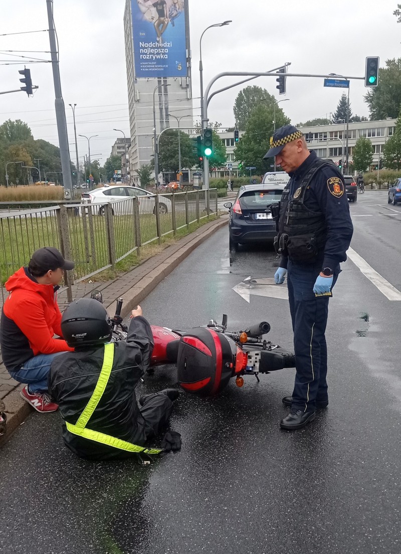 Strażnik miejski stojący przy motocykliście leżącym na jezdni. Za nimi przewrócony motocykl.