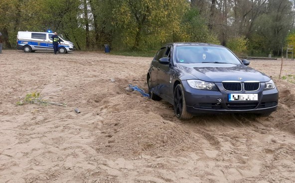 Czarne BMW zakopane w nadwiślańskim piachu. Za nim, w oddali, radiowóz straży miejskiej.