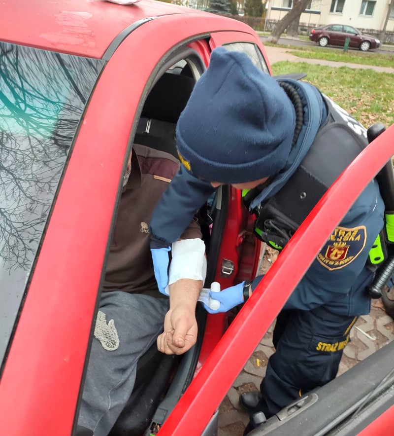 Strażniczka miejska zakładająca opatrunek na rękę mężczyźnie siedzącemu w czerwonym samochodzie.
