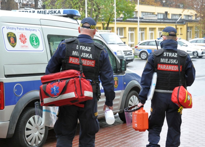 Strażnicy- ratownicy z wyposażeniem medycznym idący w stronę radiowozu z emblematami Ulicznego Patrolu Medycznego